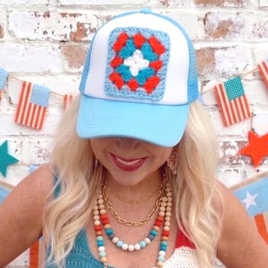 May include: A light blue trucker hat with a white mesh back and a square crochet patch in red, white, and blue. The hat is part of a patriotic theme, with a matching crochet top and beaded necklaces.
