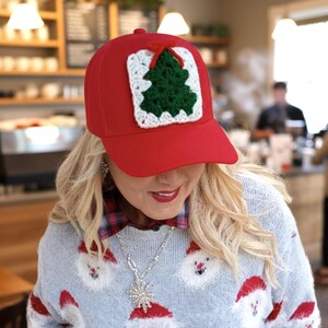 May include: A red baseball cap with a crocheted Christmas tree patch. The patch features a green tree, white border, and a red bow. The cap is worn with a light blue sweater with Santa Claus faces.