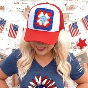 May include: A red and white trucker hat featuring a square, multi-colored crochet patch on the front panel. The patch displays a central light blue star surrounded by white and red yarn patterns. The hat has a solid red brim and a mesh back.