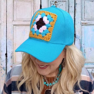 May include: A turquoise baseball cap with a square, crocheted patch. The patch features a floral design with a black center, pink and blue petals, and a gold border. The cap has a curved brim and a rounded top.