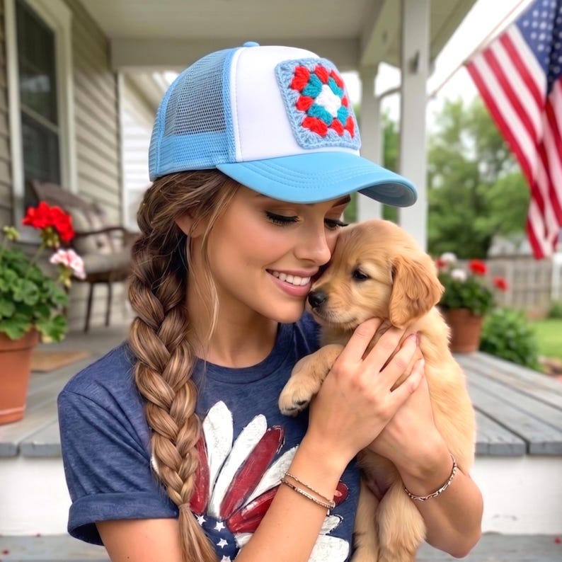 May include: A light blue and white trucker hat with a crocheted square design. The hat has a mesh back and a curved brim. The person is wearing a blue t-shirt with a red and white graphic design. A golden retriever puppy is being held.