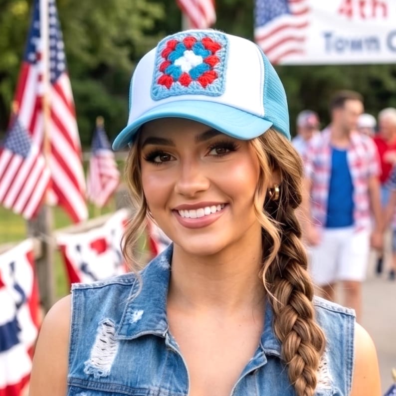 May include: A light blue and white trucker hat with a crocheted square patch in red, white, and blue. The hat has a mesh back and a curved brim. The hat is worn outdoors with American flags in the background.