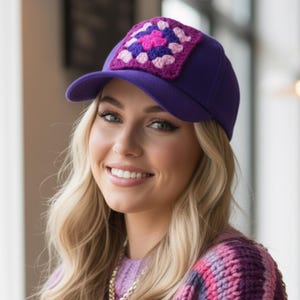 May include: A purple baseball cap with a crocheted square patch in shades of pink, purple, and white. The cap has a curved brim and a structured crown. The patch is centered on the front of the cap.
