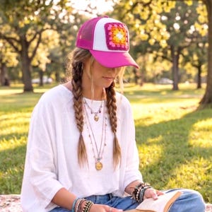 May include: A pink trucker hat with a white front panel and a crocheted square design in the center. The hat has a pink brim and mesh backing. The hat is worn outdoors in a park setting.