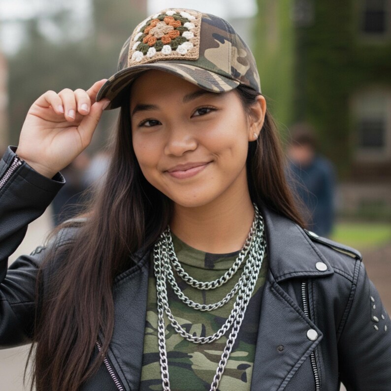 May include: Camouflage baseball cap with a square crochet patch in shades of beige, brown, and green. The cap is worn with a black leather jacket and a layered silver chain necklace. The shirt underneath is also camouflage.