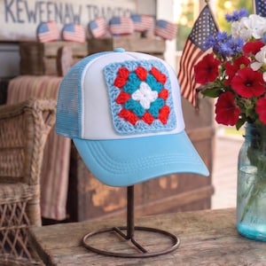 May include: A light blue and white trucker hat with a crocheted square in red, white, and blue. The hat is displayed on a metal stand. The background includes American flags and a sign that reads "KEWEENAW TRADITIONS".