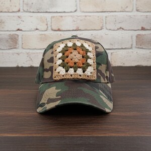 May include: Camouflage baseball cap with a crocheted square patch. The patch features a granny square design with cream, olive green, and orange colors. The cap is made of a durable fabric and has a curved brim.