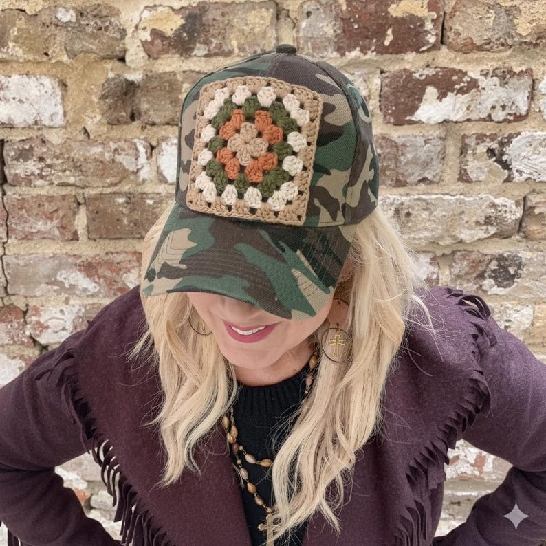 May include: Camouflage baseball cap with a crocheted square patch in shades of beige, green, and orange. The cap features a classic six-panel design and a curved brim. The hat is made of a durable fabric and is suitable for outdoor activities.
