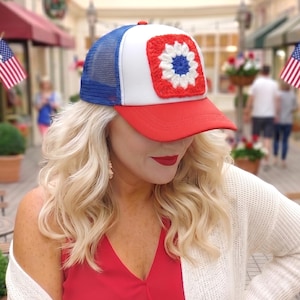 May include: A trucker hat featuring a white front panel with a square red, white, and blue crochet flower patch. The hat has a red brim, a blue mesh back, and a blue top button. The design incorporates a patriotic color scheme of red, white, and blue.