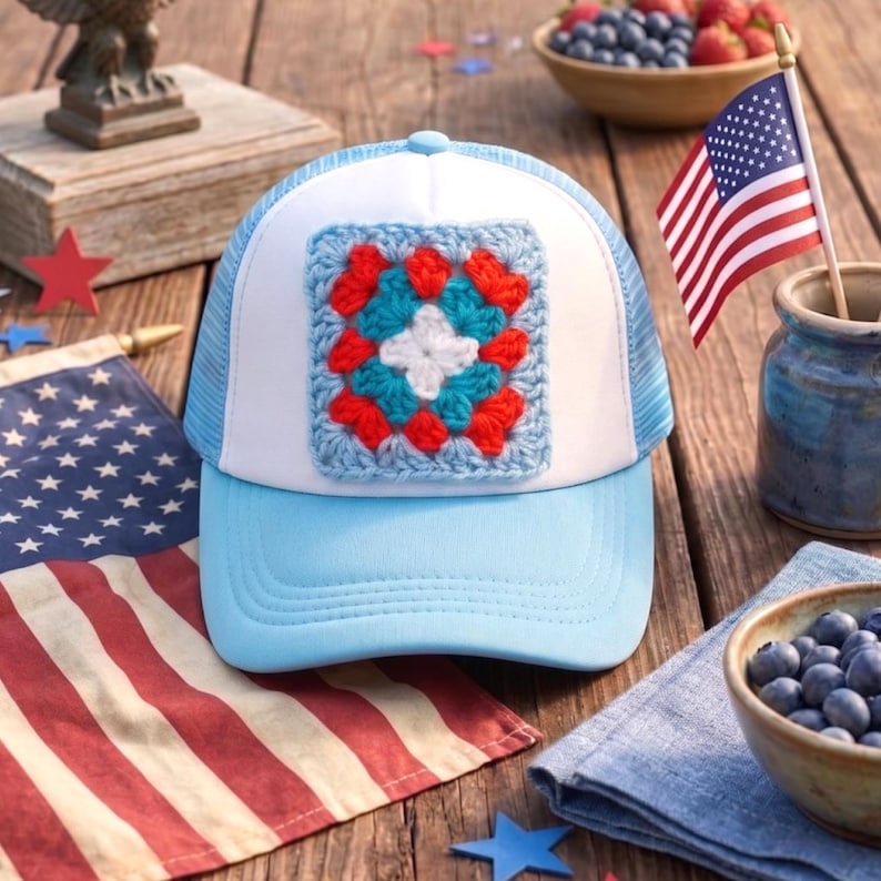 May include: A light blue and white trucker hat with a crocheted square design in red, white, and blue. The hat has a mesh back and a curved brim. The image also includes an American flag, blueberries, and strawberries.