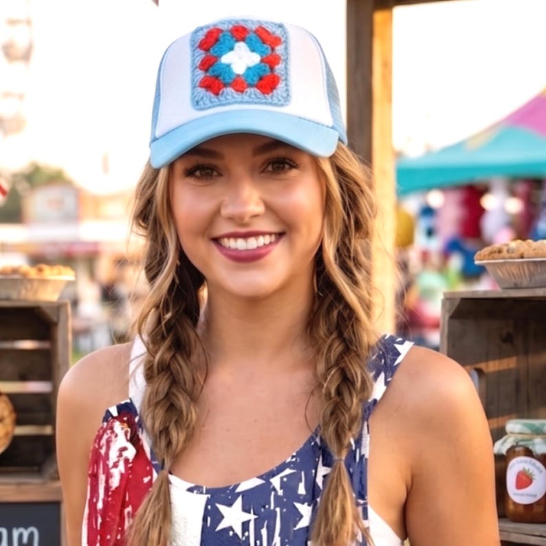 May include: A light blue and white trucker hat with a crocheted square design in red, white, and blue. The hat has a mesh back and a curved brim. The hat is a patriotic accessory.