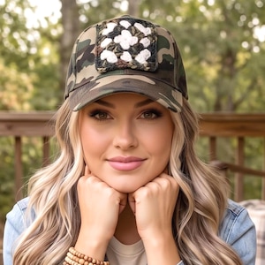 May include: A camouflage baseball cap featuring a square, crocheted granny square patch on the front panel. The patch is composed of white, dark green, and brown yarn in a floral-inspired pattern, centered on the crown of the cap.