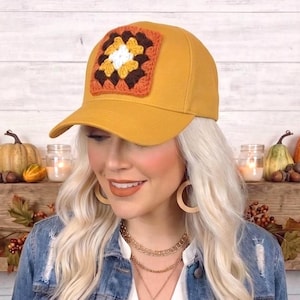 May include: A mustard yellow baseball cap featuring a square crochet granny square patch on the front. The patch is composed of concentric layers in white, yellow, and dark brown yarn, stitched onto the center of the cap's crown.