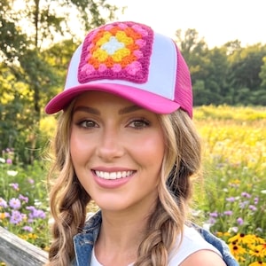 May include: A pink and white trucker hat with a crocheted square design in the center. The crocheted square features a floral pattern with shades of pink, orange, yellow, and white. The hat has a pink brim and a white mesh back.