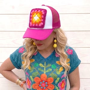 May include: A pink and white trucker hat with a square, colorful crochet patch. The hat has a pink brim and mesh back. The woman in the image is wearing a teal embroidered shirt and yellow earrings.