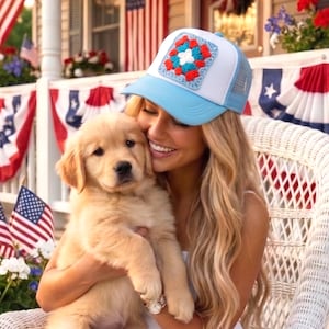 May include: A light blue and white trucker hat with a crocheted square design in red, white, and blue. The hat is worn by a person holding a golden retriever puppy. The background includes American flags and patriotic decorations.