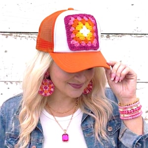 May include: An orange and white trucker hat with a square, multi-colored crochet patch. The hat has a mesh back and a curved brim. The crochet patch features shades of pink, orange, yellow, and white. The hat is a stylish accessory.