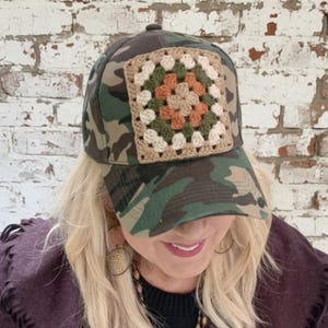 May include: Camouflage baseball cap with a square, crocheted patch. The patch features a granny square design in beige, green, and orange. The cap is a classic six-panel design with a curved brim.