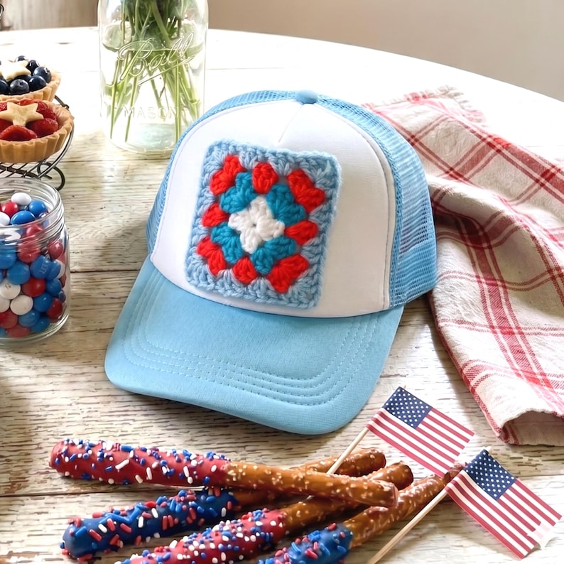 May include: A light blue and white trucker hat with a crocheted square patch in red, white, and blue. The hat is displayed with patriotic treats and small American flags, perfect for a summer celebration.