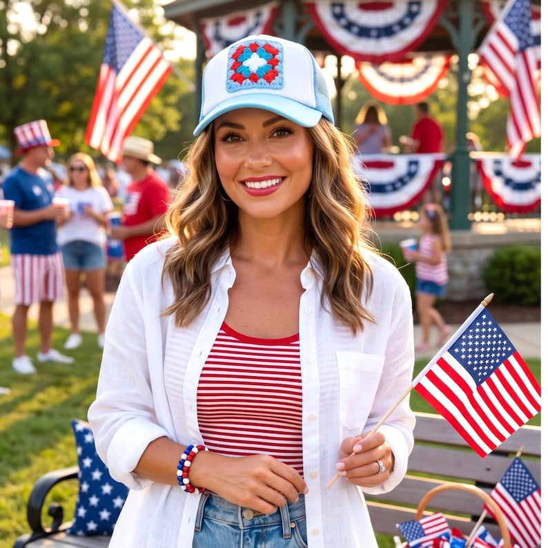 May include: A woman wearing a white button-down shirt over a red and white striped tank top, and a light blue and white trucker hat with a crochet patch. She is holding a small American flag.