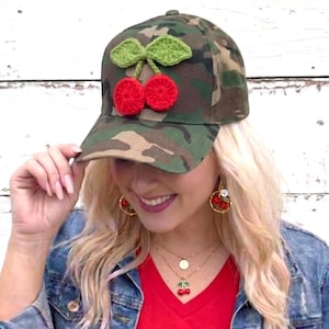 May include: Camouflage baseball cap with a crocheted cherry design. The cap features two red cherries and green leaves. The hat is a classic six-panel design with a curved brim.