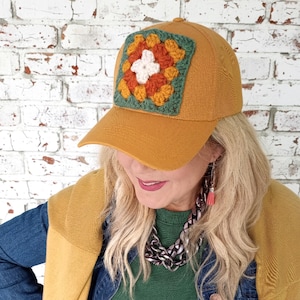 May include: Mustard yellow baseball cap with a square, crocheted patch. The patch features a white flower in the center, surrounded by orange and yellow petals, all framed by a green border. The cap is a classic six-panel design.