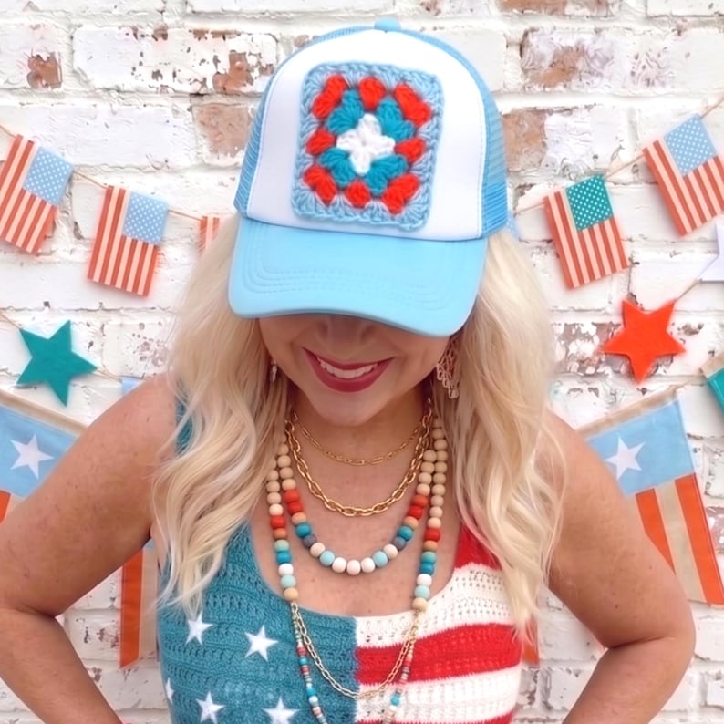 May include: A light blue trucker hat with a white mesh back and a square crochet patch in red, white, and blue. The hat is part of a patriotic theme, with a matching crochet top and beaded necklaces.