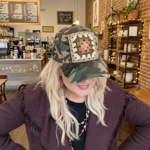 May include: Camouflage baseball cap with a square, crocheted patch in shades of beige, orange, and green. The cap features a classic six-panel design and a curved brim. The hat is a stylish accessory.