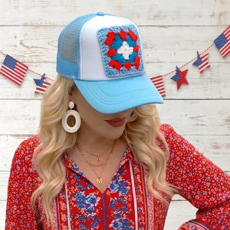 May include: A light blue trucker hat with a white front panel and a crocheted square design in red, white, and blue. The hat has a mesh back and a curved brim. The image also shows American flag decorations in the background.