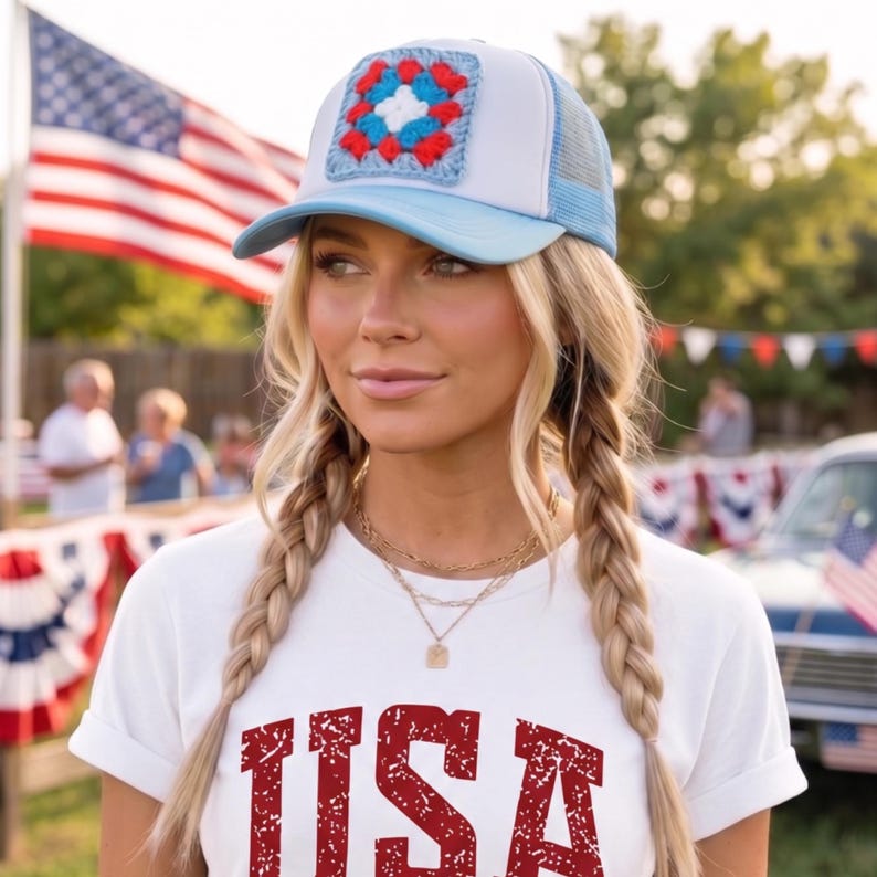 May include: A light blue and white trucker hat with a crocheted square design in red, white, and blue. The hat is worn with a white t-shirt with the word "USA" in red block letters. The background includes an American flag and festive decorations.