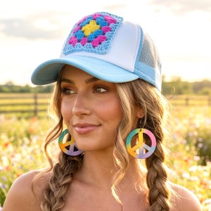 May include: A light blue and white trucker hat featuring a square, multi-colored crochet granny square patch on the front panel. The hat is paired with round, flat earrings displaying a gradient peace sign design in yellow, pink, and blue.