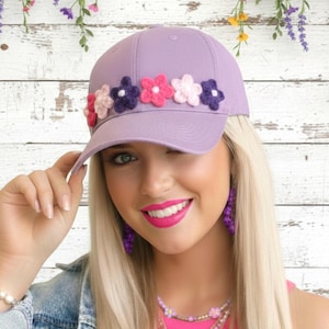 May include: A lavender baseball cap adorned with colorful, crocheted flowers. The cap features a curved brim and a classic six-panel design. The flowers are in shades of pink, purple, and white, adding a touch of whimsy to the accessory.