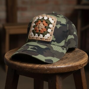 May include: A camouflage baseball cap with a crocheted square patch. The patch features a square design with a tan border and a center of orange, green, and white. The cap is sitting on a wooden stool.