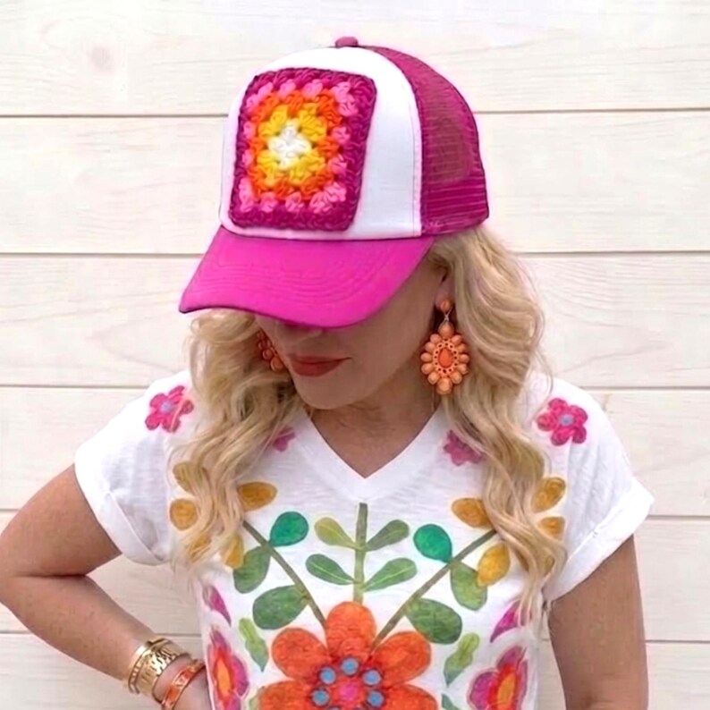 May include: A pink and white trucker hat with a square, colorful crochet patch. The hat is paired with a white V-neck t-shirt featuring a floral design in orange, green, and pink. The person is also wearing orange earrings and bracelets.