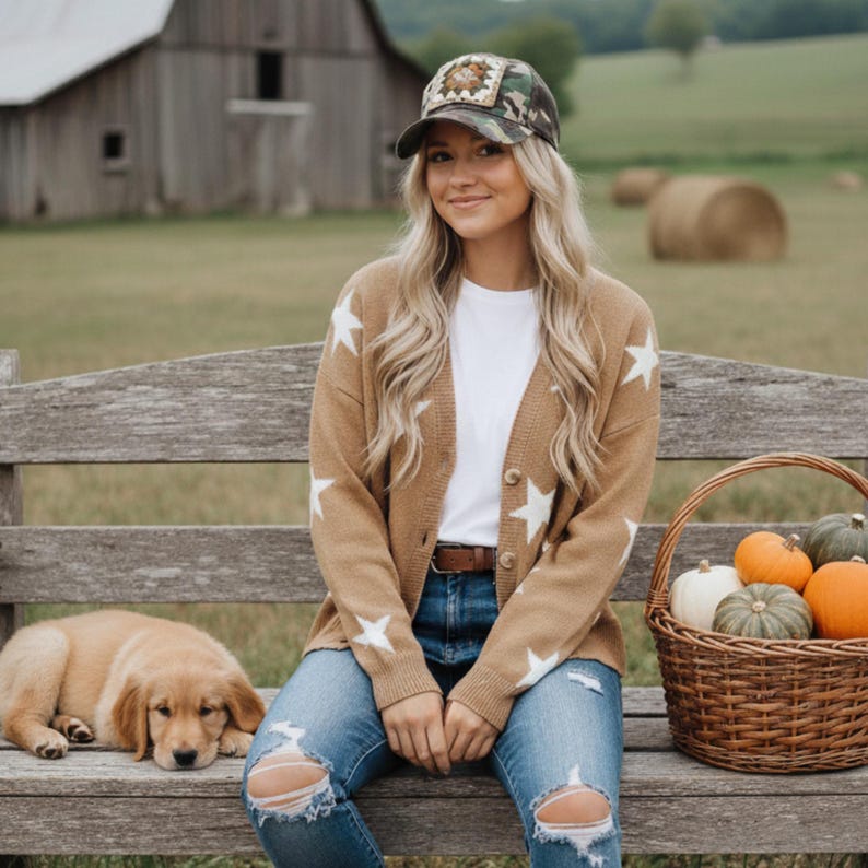 May include: A tan cardigan with white star patterns, paired with ripped blue jeans and a camouflage baseball cap. A basket of pumpkins and a sleeping puppy are also in the image, creating a cozy, autumnal scene.
