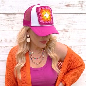 May include: A pink trucker hat with a white front panel and a crocheted square design in shades of pink, orange, yellow, and white. The hat has a mesh back and a curved brim.