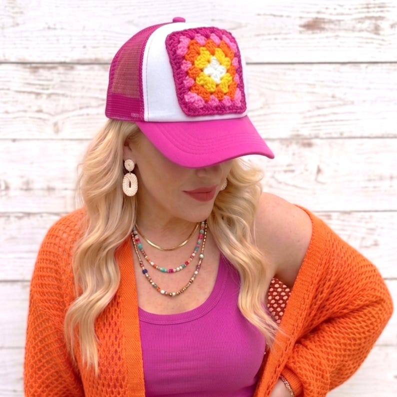 May include: A pink trucker hat with a white front panel and a crocheted square design in shades of pink, orange, yellow, and white. The hat has a mesh back and a curved brim.