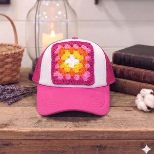 May include: A pink trucker hat with a white front panel and a crocheted square design in the center. The square features a floral pattern with pink, orange, yellow, and white colors. The hat has a pink brim and mesh back.