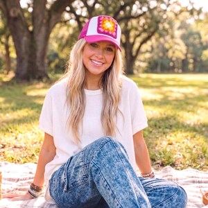 May include: A pink and white trucker hat with a square, crocheted patch in shades of yellow, pink, and orange. The hat is worn by a person sitting on a blanket in a park setting. The person is wearing a white shirt and blue jeans.
