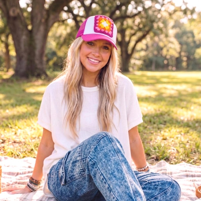 May include: A pink and white trucker hat with a square, crocheted patch in shades of yellow, pink, and orange. The hat is worn by a person sitting on a blanket in a park setting. The person is wearing a white shirt and blue jeans.