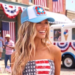 May include: A light blue and white trucker hat with a crocheted square patch in red, white, and blue. The hat has a mesh back and a curved brim. The person is wearing a strapless top with a red, white, and blue American flag design.