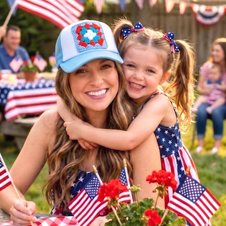 May include: A woman and a young girl embrace, both smiling. The woman wears a light blue trucker hat with a crocheted square. The girl wears a dress with a star and stripe pattern. Red flowers and American flags are in the foreground, with a festive outdoor setting.