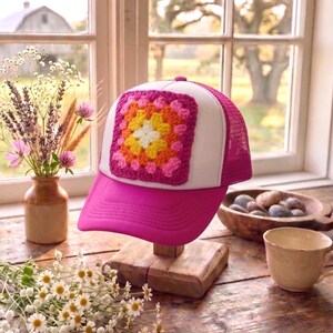 May include: A pink trucker hat with a white front panel and a pink mesh back. The hat features a square, colorful crochet patch in the center. The crochet patch has a floral design with pink, orange, yellow, and white colors.