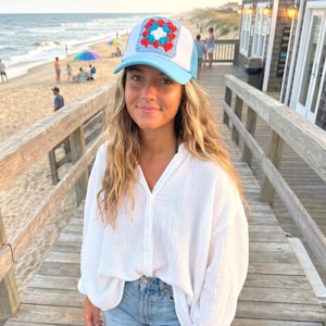 May include: A light blue and white trucker hat with a square, multi-colored crochet patch. The hat has a mesh back and a curved brim. The crochet patch features red, white, and blue squares. The hat is worn outdoors.