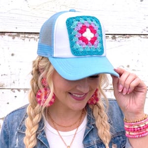 May include: A light blue trucker hat with a white front panel and a crocheted square patch. The patch features a colorful granny square design in shades of turquoise, pink, purple, and white. The hat has a curved brim and a mesh back.