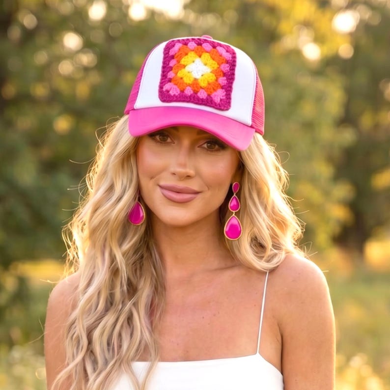 May include: A pink and white trucker hat with a crocheted square design in the center. The square features a colorful pattern of yellow, orange, and pink. The hat has a pink brim and a white mesh back.