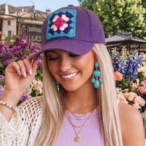 May include: A purple baseball cap with a square, crocheted patch in shades of pink, blue, and white. The cap is worn by a person with blonde hair, turquoise earrings, and layered gold necklaces. The background features flowers and a sign that reads "Lilacs."