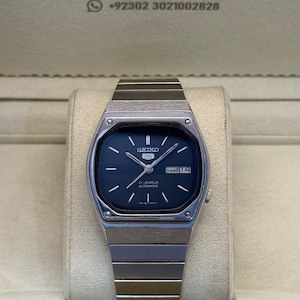 May include: A silver-toned Seiko 5 automatic wristwatch with a dark blue dial, silver hour markers, and a metal link bracelet. The watch face displays the Seiko logo, 21 jewels text, and a day-date window. The watch is presented in a branded box.