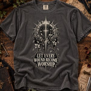 May include: A charcoal grey short-sleeve t-shirt featuring a graphic of a cracked circular stone frame surrounding a cross with radiating light and floral vines. The text "LET EVERY WOUND BECOME WORSHIP" is printed in white below the graphic.