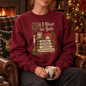 Może przedstawiać: Ciemnoczerwona bluza z okrągłym dekoltem z grafiką kota śpiącego na stosie książek. Tekst brzmi All I Want for Yule, Good Books, Warm Blankets, Hot Drinks, And No Responsibilities, w towarzystwie zapalonej świecy i gałązek ostrokrzewu.